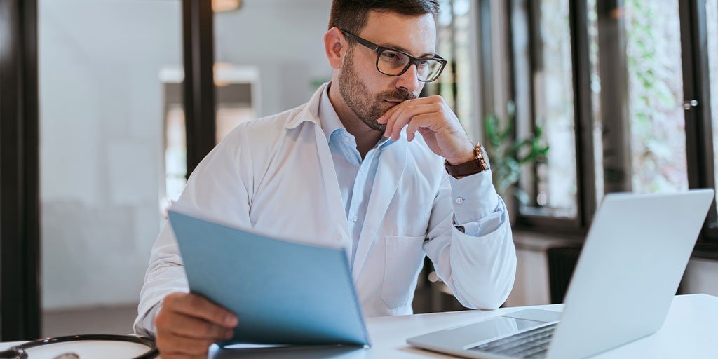 Doctor on laptop with documents in other hand.